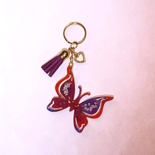 Portachiavi farfalla Lunéa in resina arancione e viola con dettagli floreali, charm dorati e nappina porpora.