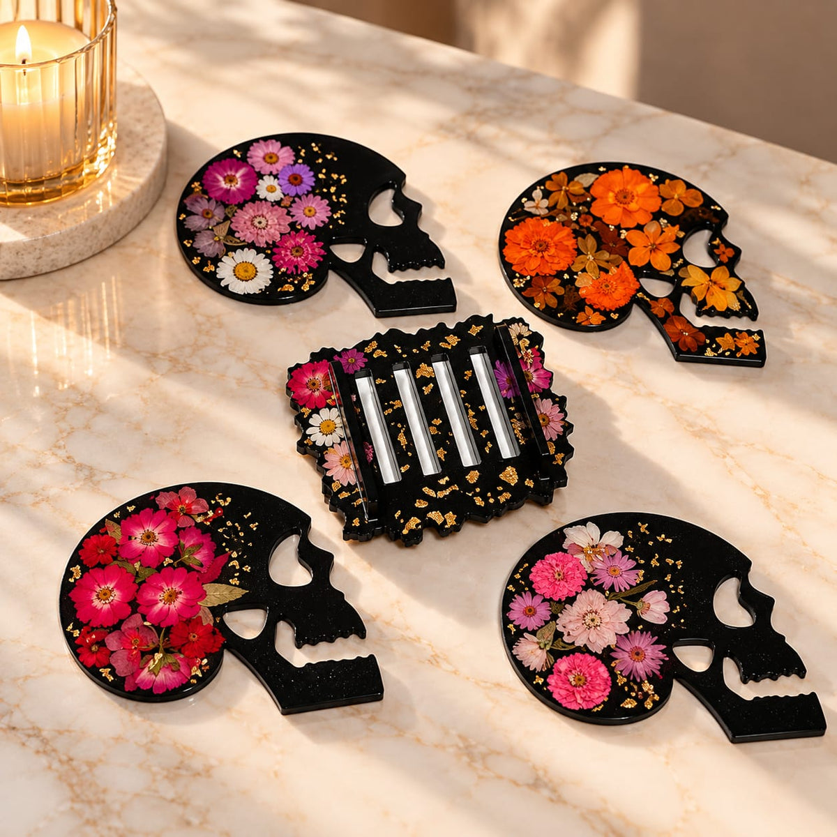 Quattro sottobicchieri artigianali a forma di teschio in resina nera, decorati con fiori pressati rosa, fucsia e arancioni, accompagnati dal supporto coordinato con dettagli dorati.